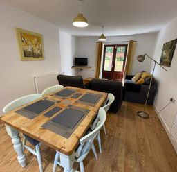 A dining table with four chairs and placemats in a room with sofas a floor lamp and a television at Stable Cottage in Shipham