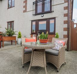 An outdoor patio area with a round wicker table and two wicker chairs with cushions and pillows in front of a house with potted plants and a wooden gate at Bwlch Y Gwynt Bach in Llangefni