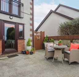 A patio with a round table and two chairs with cushions and plants in pots near a house at Bwlch Y Gwynt Bach in Llangefni