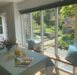 A dining area with a table covered in a blue polka dot tablecloth and windows with a garden view at Ribby Farmhouse in Lerryn