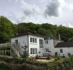 A white two-story house with multiple windows and a wooden deck surrounded by trees and greenery at Ribby Farmhouse in Lerryn