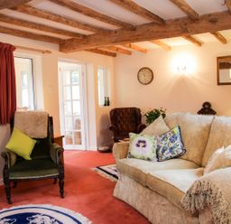 A living room with a beige sofa and floral cushions a green armchair and wooden ceiling beams at Cream Door Cottage in Kidderminster