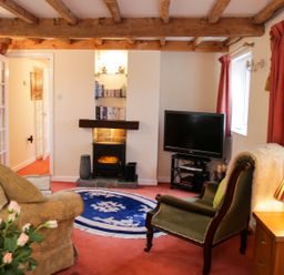 A living room with wooden beams on the ceiling a TV a fireplace an armchair a sofa a round blue rug and flowers on a table at Cream Door Cottage in Kidderminster