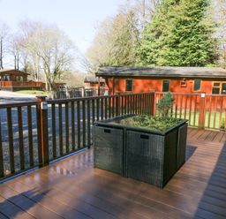 An outdoor wooden deck with a rattan table and railing overlooking wooden cabins and trees at 40 Skiptory Howe in Troutbeck Bridge