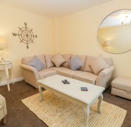 A living room with a beige sofa set and cushions a white coffee table with remotes and a coaster a side table with a lamp and decor a round wall mirror and a compass wall art at Valiant House in Scarborough