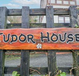 A wooden sign with Tudor House written on it attached to a black wooden fence with a house in the background at Tudor House in Symonds Yat