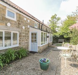 A stone exterior of a house with white framed windows a white door a small patio area with a white metal table and chairs and garden plants at Robin Cottage in Morpeth