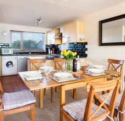 A dining area with a wooden table set for six and an open kitchen with appliances at 164 Cumber Close in Malborough