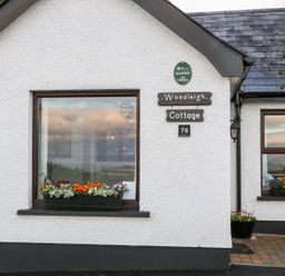 The exterior of Woodleigh Cottage with windows displaying flower boxes and a sign with the cottage name and number 76 at Ballycastle