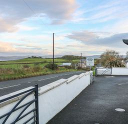 A driveway and paved area next to a white wall and gate with a road and green fields in the background at Woodleigh Cottage in Ballycastle
