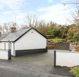 A driveway with a white house on the left and steps leading to a garden with greenery and a picnic table at Woodleigh Cottage in Ballycastle