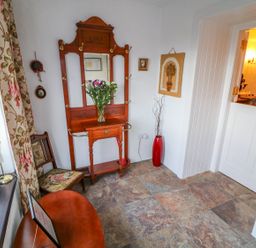 A small room with a wooden hall stand with a mirror a chair a red vase with branches and a wooden table by a window at Woodleigh Cottage in Ballycastle