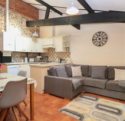 An open kitchen and living area with a dining table with chairs a gray sectional sofa and a wall clock at Hitchens Cottage in Happisburgh