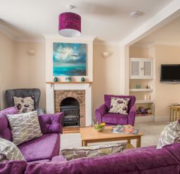 A living room with purple sofas and armchairs a fireplace with painting above and a wall-mounted television at Wey Cottage in Weymouth
