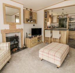 A living room with a plaid armchair and ottoman a TV on a wooden stand a fireplace and an open kitchen with bar stools at 27 Lake View in Cawston