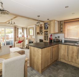 An open plan kitchen and living area with wooden cabinets dining table armchair and large window blinds at 27 Lake View in Cawston