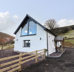A white house with a triangular roof and large windows surrounded by a wooden fence in a rural setting at Tyn Llwyn Barn in Glyndyfrdwy near Llangollen