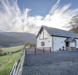 A white barn with a slate roof and a fenced gravel driveway on a hillside with mountains in the background at Tyn Llwyn Barn in Glyndyfrdwy near Llangollen
