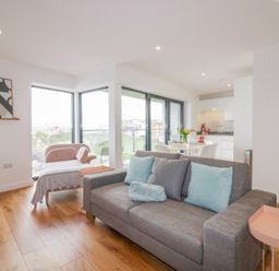 A living room with a gray sofa and pink side table and a small dining area and kitchen at Onshore 7 in Newquay