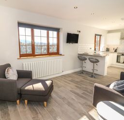 A living room with two grey armchairs and an ottoman near a window and an open kitchen with a breakfast bar and stools at Afon Rhos Bach in Llanrug