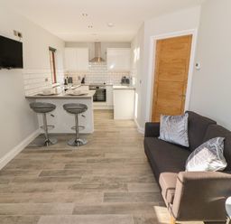 A kitchen with a breakfast bar and two stools next to a living area with a sofa and cushions at Afon Rhos Bach in Llanrug