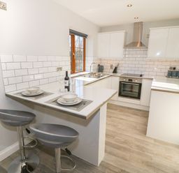 A kitchen with white cabinets a stove a sink and two gray bar stools at Afon Rhos Bach in Llanrug