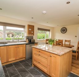 A kitchen with a wooden island, dining table with chairs, and windows overlooking greenery at New Peny in Llandrindod Wells