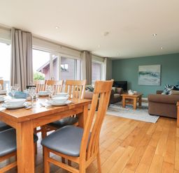 A dining table with wooden chairs and tableware next to a living room with sofas and a coffee table at Lodge Sixty Six in St Columb Major