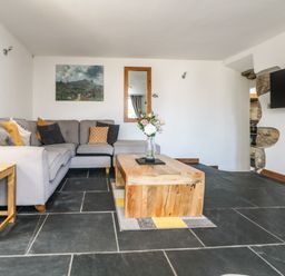 A living room with a grey sectional sofa wooden coffee table with flowers a wall-mounted television and stone flooring at Miner's Rest in Troon near Camborne