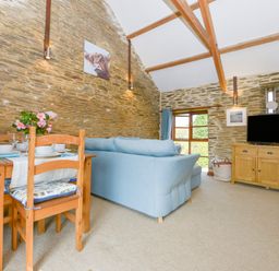 A living room with a wooden dining table and chairs a blue sofa a wooden cabinet with a television and stone walls at Erin Cottage in Pelynt near Looe