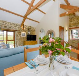 A dining table with flowers and place settings overlooking a living room with a blue sofa and exposed stone walls at Erin Cottage in Pelynt near Looe