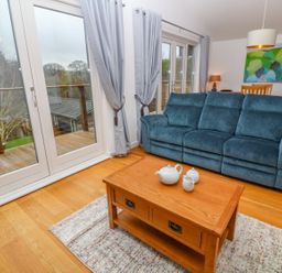 A living room with a blue sofa a wooden coffee table with a tea set and glass doors with curtains at 1 Lake View in Lanreath