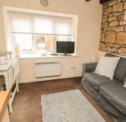 A small living room with a gray sofa and cushions a dining table with tea set a window with a TV on the sill and a stone wall at Wren Cottage in Morpeth