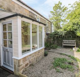 A stone house with a white door and windows next to a garden with a wooden bench and plants at Wren Cottage in Morpeth