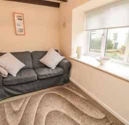 A small living room with a gray sofa two pillows a patterned rug and a windowsill with a lamp bowl and vase of yellow flowers at Heron Cottage in Morpeth