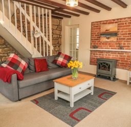 A living room with a gray sofa with red and checkered cushions a wooden coffee table with yellow flowers a brick fireplace and a white door at Magpie Cottage in Morpeth