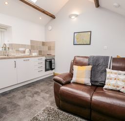 A kitchen area with white cabinets and a brown leather sofa with cushions at La'al Riddings in Lowick Green Riddings Farm Near Ulverston