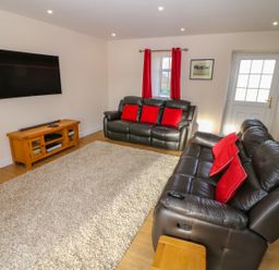 A living room with two black leather sofas with red cushions a wooden TV stand with a flat screen TV mounted on the wall and a beige rug at Stabl in Pwllheli