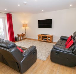 A living room with two black leather sofas red cushions a beige rug curtains a wooden tv stand with a mounted flat screen tv and a floor lamp at Stabl in Pwllheli
