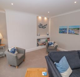 A living room with grey armchairs, a blue sofa, a wooden side table with a lamp, wall art, and built-in shelves at Moorings in Kingswear Devon