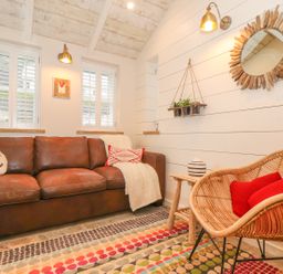 A living room with a brown leather sofa two pillows a wicker chair with two red pillows a wooden side table with a vase a round wooden mirror on the wall and hanging plants at Beach Hut in Trebarwith Strand