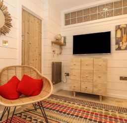 A room with a wicker chair and red cushions a patterned rug a wooden cabinet a wall-mounted TV a small shelf with a globe a wooden door and a round mirror at Beach Hut in Trebarwith Strand