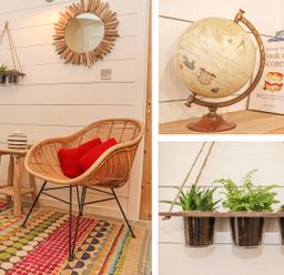 A corner of a room with a wicker chair with red cushions a wooden stool with a vase a hanging planter with three potted plants a round sunburst mirror and a globe with books on a shelf at Beach Hut in Trebarwith Strand