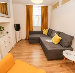 A living room with a gray sectional sofa with yellow cushions a yellow armchair a white round table wooden flooring and mustard curtains at West Cliff Apartment in Whitby