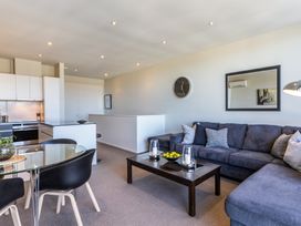 An open-plan living room and kitchen with a glass dining table, black chairs, gray sectional sofa, coffee table, wall clock, and wall mirror at 24 Marina View Retreat Taupo Holiday Apartment Taupo Bay