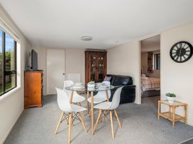 A dining area with a round glass table surrounded by white chairs a wooden cabinet a black sofa a wall clock and a small table with a plant in Acacia Views - Acacia Bay Holiday Home in Acacia Bay