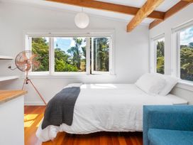 A bedroom with a bed white pillows and blanket a gray throw a blue chair a standing fan wooden beams on ceiling and large windows showing greenery at Piha City Escape Piha Holiday Home in Piha