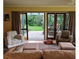 A living room with patterned armchairs and a sofa facing glass doors opening to a wooden deck and green garden at Surf and Sun Retreat - Mt Maunganui Holiday Home in Mt Maunganui