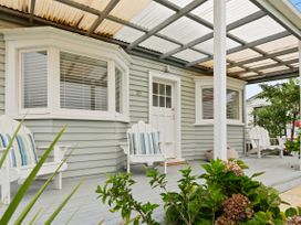 A porch with white wooden chairs and a striped cushion outside a house with light green siding and a corrugated plastic roof at The Wallnutt Nook - Waihi Beach