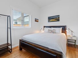 A bedroom with a bed with white bedding a nightstand with a lamp a window with blinds and a framed picture on the wall at The Wallnutt Nook - Waihi Beach Holiday Home in Waihi Beach